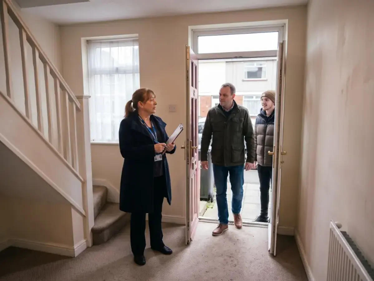 A housing officer with a clipboard greets two visitors at the open front door of a home
